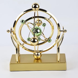 Könnte beinhalten: Eine goldfarbene kinetische Kunstskulptur mit Sockel und Stützrahmen. Die Skulptur zeigt mehrere konzentrische Ringe und Kugeln in Gold und Grün, die ein dynamisches, orbitales Design bilden. Der Sockel ist rechteckig und ebenfalls goldfarben.