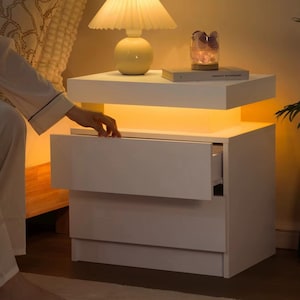 May include: A modern white nightstand with two drawers, one partially open. The nightstand has a floating shelf with a warm light underneath. A lamp and decorative items sit on top. The nightstand is next to a bed.