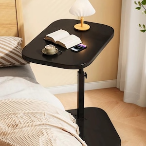 May include: A black adjustable bedside table with a rectangular top. A small lamp, an open book, a smartphone, and a cup of coffee sit on the table. The table is positioned next to a bed with a patterned pillow and a beige blanket.