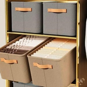 Peut inclure: Unité de rangement à plusieurs niveaux avec des bacs en tissu gris et beige. Les bacs ont des poignées en similicuir beige. Les étagères sont remplies de vêtements pliés. Le cadre est doré.