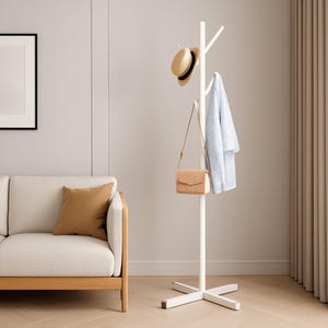 May include: A white coat stand with a cross-shaped base. A straw hat with a black band, a light blue garment, and a tan handbag hang from the stand. The stand is in a room with a beige sofa and a brown cushion.