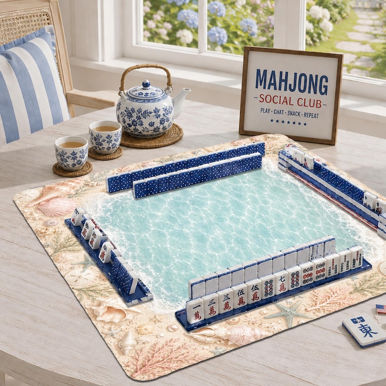 Op de afbeelding: Een mahjongset met witte tegels met blauwe en rode karakters, gerangschikt op een rechthoekige speelmat met een strandprint van zand, schelpen en turquoise water. Een blauw-witte theepot met bloemmotief en twee bijpassende kopjes staan ernaast.