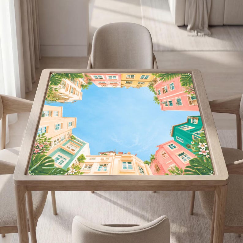 Puede incluir: Una mesa cuadrada de madera con un estampado en el tablero de edificios coloridos en tonos rosa, amarillo y turquesa que rodean un cielo azul brillante. El dise&ntilde;o incluye follaje tropical verde y flores blancas que enmarcan la arquitectura.