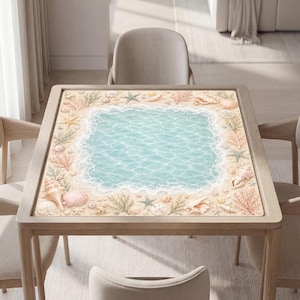 Op de afbeelding: Een vierkante tafel met een bedrukt tafelblad met een lichtblauw oceaancentrum, omringd door een zandkleurige rand vol diverse schelpen, zeesterren en koraal in tinten beige, roze en zachtgroen, rustend op vier houten poten.