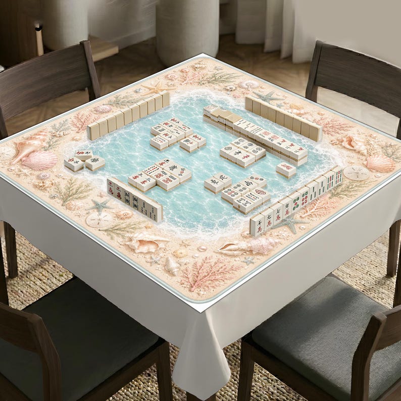 Op de afbeelding: Een vierkant mahjong-tafelkleed met een strandthema, voorzien van lichtblauw water, wit zand, schelpen en koraal. Op het oppervlak liggen ivoorkleurige mahjong-tegels met diverse zwarte en rode Chinese karakters en symbolen afgebeeld.