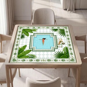 May include: A square table featuring a printed surface with a light blue swimming pool graphic, a person floating in the water, and another person on a lounge chair. The design includes green palm leaves and a white and light gray checkered pattern.