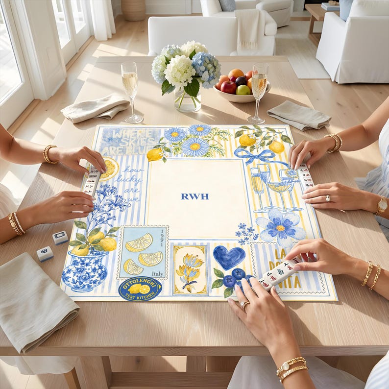 May include: A square mahjong mat featuring a blue and yellow watercolor design with lemons, flowers, and the text "WHERE SKIES ARE BLUE", "how lucky are we", "RWH", "1991", "Italy", and "OTTOLENGHI TEST KITCHEN" printed on a light background.