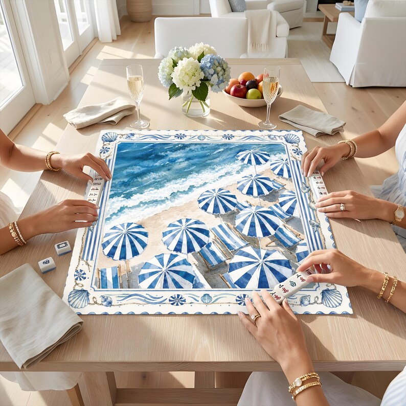 May include: A square game board featuring a coastal scene with blue and white striped beach umbrellas and lounge chairs on a sandy shore. The border is decorated with blue seashells and rope patterns. Small game tiles are placed on the board.