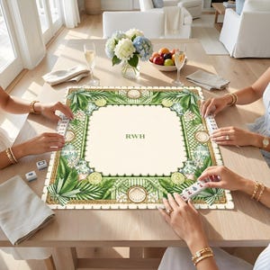 K&ouml;nnte beinhalten: Eine quadratische Mahjong-Matte mit cremefarbener Mitte und den gr&uuml;nen Buchstaben RWH. Der Rand zeigt ein gr&uuml;nes botanisches Muster mit Palmbl&auml;ttern, Zitrusfr&uuml;chten und Muscheln. Die Matte liegt auf einem Holztisch mit Spielsteinen.