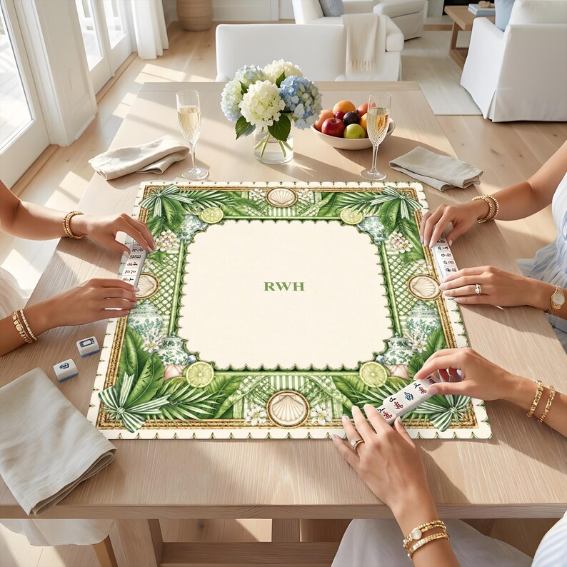 K&ouml;nnte beinhalten: Eine quadratische Mahjong-Matte mit cremefarbener Mitte und den gr&uuml;nen Buchstaben RWH. Der Rand zeigt ein gr&uuml;nes botanisches Muster mit Palmbl&auml;ttern, Zitrusfr&uuml;chten und Muscheln. Die Matte liegt auf einem Holztisch mit Spielsteinen.