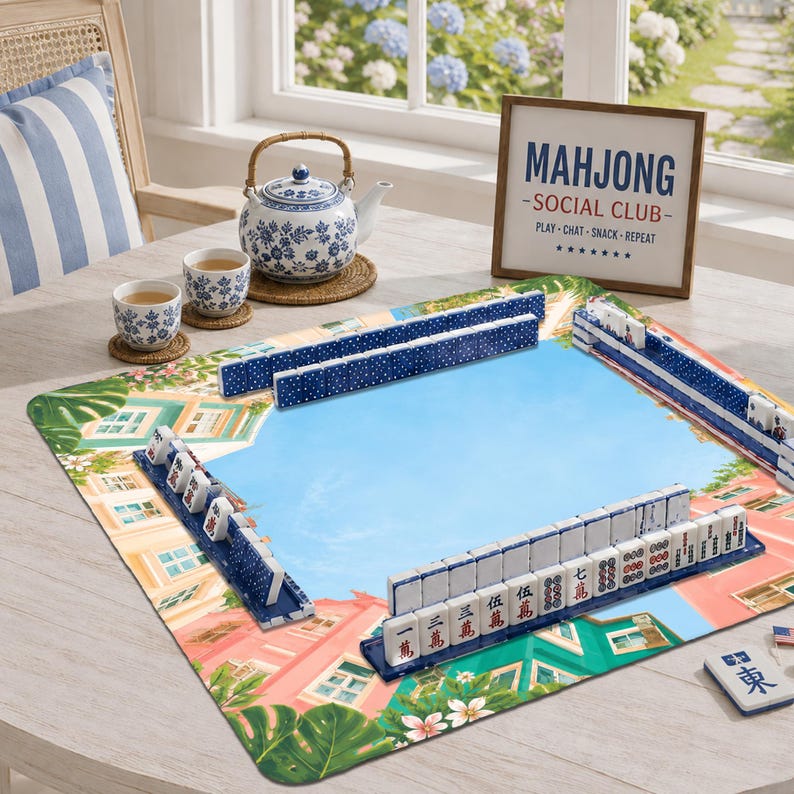 Puede incluir: Un juego de Mahjong con fichas blancas con caracteres rojos y negros sobre una alfombrilla de juego colorida con estampado de edificios tropicales. Una tetera de cer&aacute;mica azul y blanca y dos tazas a juego, junto a un letrero enmarcado que dice MAHJONG SOCIAL CLUB PLAY-CHAT-SNACK-REPEAT.