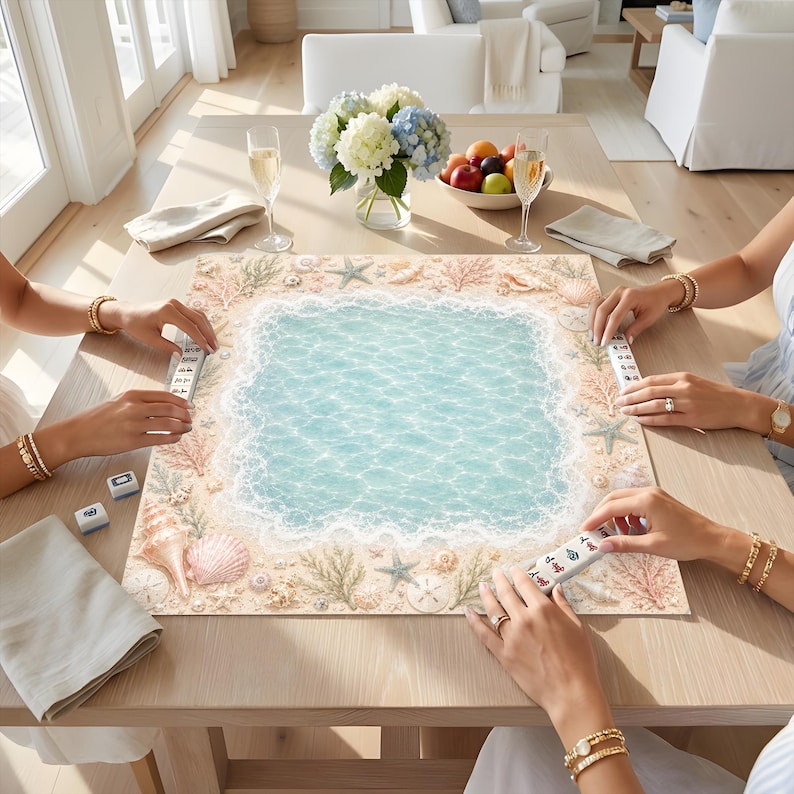 Op de afbeelding: Een vierkante mahjongmat met een centraal lichtblauw waterontwerp met witte schuimranden, omringd door een rand van beige zand, schelpen, zeesterren en koraal. Vier witte mahjongtegels met zwarte en rode karakters liggen op de mat.
