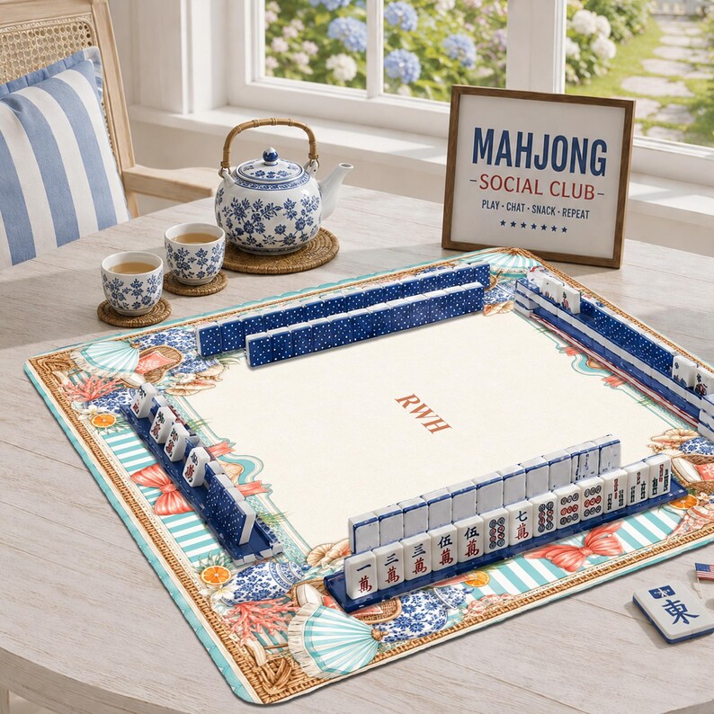 May include: A square mahjong mat with a cream center featuring the initials RWH, surrounded by a border of blue and white patterns, coral, and citrus motifs. Blue tile racks hold white mahjong tiles with red and black characters.