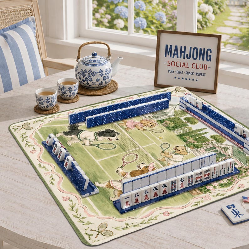 May include: A mahjong set featuring blue and white tiles arranged on a green mat with a tennis-themed illustration of dogs. A blue and white floral teapot and two matching cups sit nearby, alongside a sign reading MAHJONG SOCIAL CLUB.