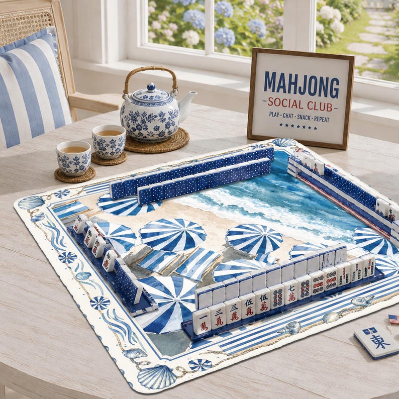May include: A square mahjong mat featuring a beach scene with blue and white umbrellas, accompanied by a blue and white floral teapot, two matching teacups, and a framed sign that reads MAHJONG SOCIAL CLUB PLAY-CHAT-SNACK-REPEAT.