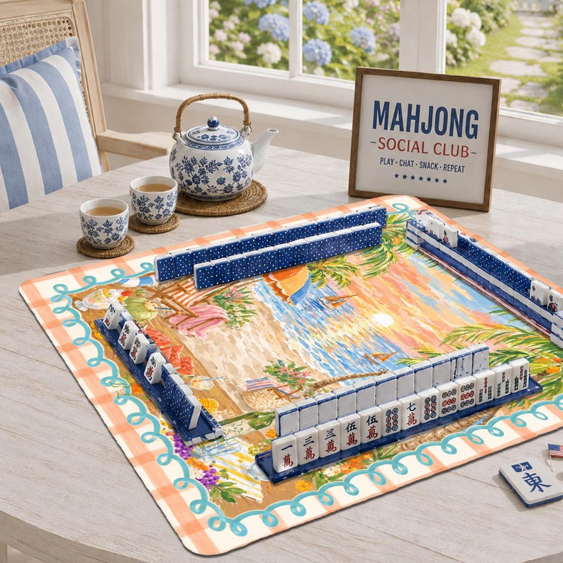 Op de afbeelding: Een mahjongset met witte tegels met rode en zwarte karakters, gerangschikt op een vierkante mat met een strandprint. Inclusief een blauw-witte theepot, twee bijpassende kopjes en een ingelijst bord met de tekst MAHJONG SOCIAL CLUB.