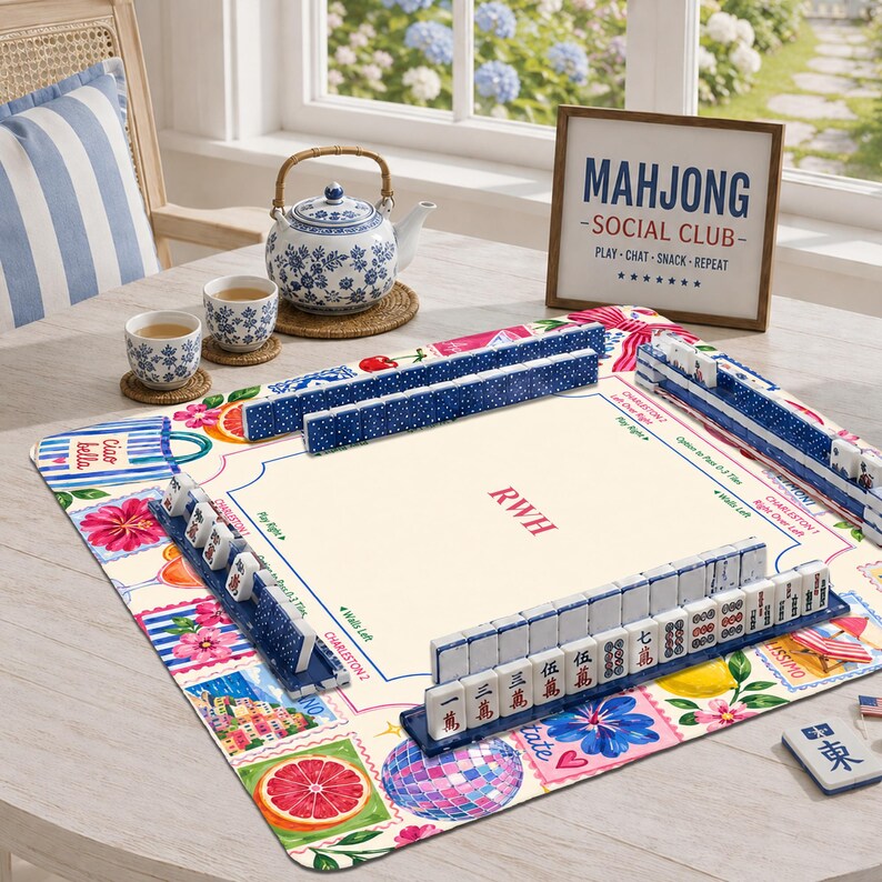 May include: A mahjong set featuring a cream-colored play mat with colorful floral and fruit patterns, blue tile racks, and white tiles with black and red characters. A blue and white porcelain teapot and two matching teacups sit nearby.