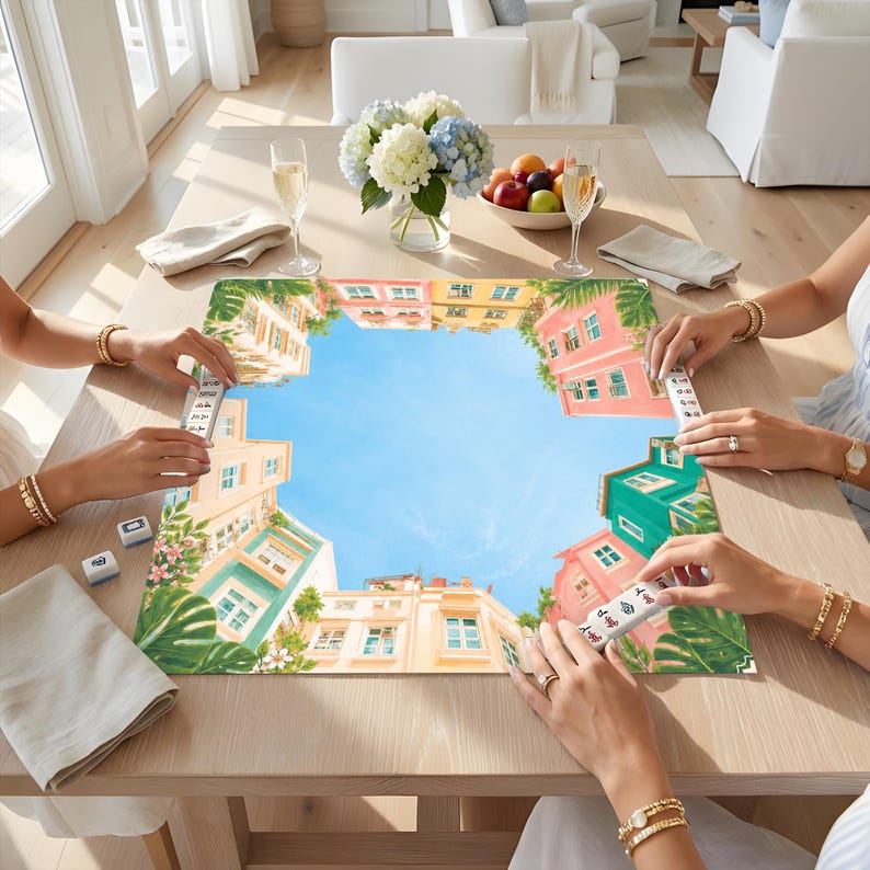 Puede incluir: Un tapete de mahjong cuadrado con una ilustraci&oacute;n colorida de edificios de estilo mediterr&aacute;neo que rodean un cielo azul brillante, con hojas tropicales verdes en las esquinas, colocado sobre una mesa de madera con fichas y vasos.