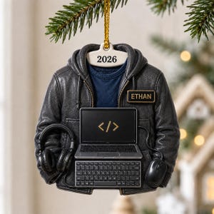 Op de afbeelding: Een hangend ornament in de vorm van een donkergrijze jas met capuchon over een marineblauw shirt. De jas heeft een naamplaatje met ETHAN, een laptop met een gouden codesymbool, een los toetsenbord, een computermuis en een koptelefoon. 2026 staat bovenaan.