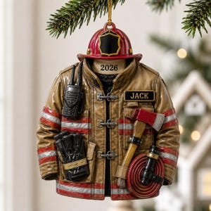 May include: A firefighter jacket ornament featuring a red helmet on top with the year 2026 printed below. The tan jacket has silver reflective stripes, a black walkie-talkie, black gloves, an axe, a coiled red hose, and a name tag reading JACK.