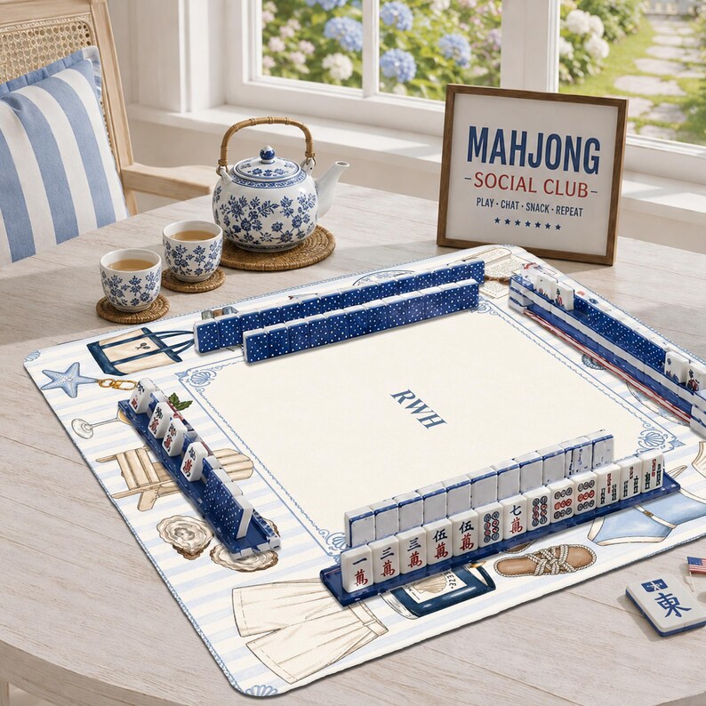 May include: A Mahjong set with blue and white tiles arranged on a patterned play mat featuring beach-themed illustrations. The mat displays the initials RWH in the center. A blue and white teapot and two matching teacups sit nearby.