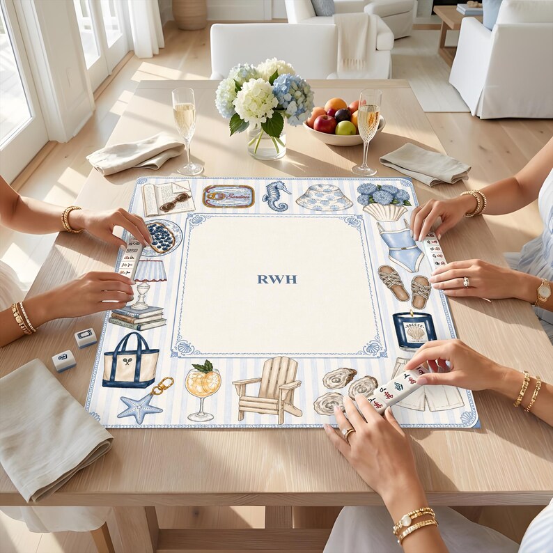 May include: A square game mat featuring a light blue and white striped border with illustrations of a tote bag, starfish, cocktail, chair, oysters, candle, sandals, swimsuit, hat, seahorse, and sardines. The center is cream with the letters RWH.