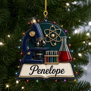 May include: A science-themed Christmas ornament featuring a dark blue microscope, a gold atom symbol, a rack with three colourful test tubes, and a red liquid-filled flask. An open book at the base displays the name Penelope in black script lettering.