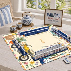 K&ouml;nnte beinhalten: Eine quadratische Mahjong-Matte mit cremefarbener Mitte und den Buchstaben RWH, umgeben von Illustrationen eines gelben Motorrads, Blumen und einer Gitarre. Blaue Fliesenhalter halten wei&szlig;e Mahjong-Steine mit roten und schwarzen Symbolen.