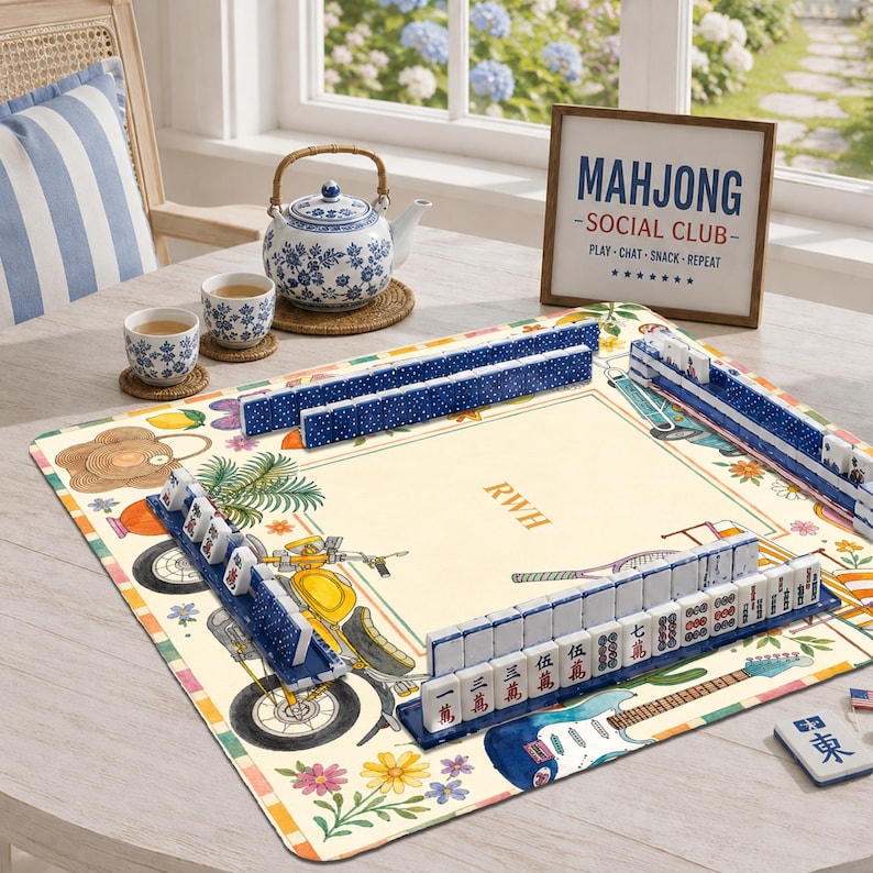 K&ouml;nnte beinhalten: Eine quadratische Mahjong-Matte mit cremefarbener Mitte und den Buchstaben RWH, umgeben von Illustrationen eines gelben Motorrads, Blumen und einer Gitarre. Blaue Fliesenhalter halten wei&szlig;e Mahjong-Steine mit roten und schwarzen Symbolen.