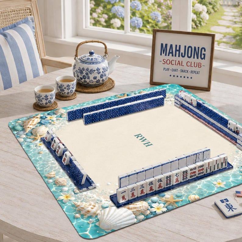 Puede incluir: Un tapete cuadrado de mahjong con un borde tem&aacute;tico de playa que presenta conchas marinas y flores blancas sobre un fondo de agua turquesa. El centro es de color crema con las letras RWH. Fichas azules de mahjong est&aacute;n dispuestas en soportes.