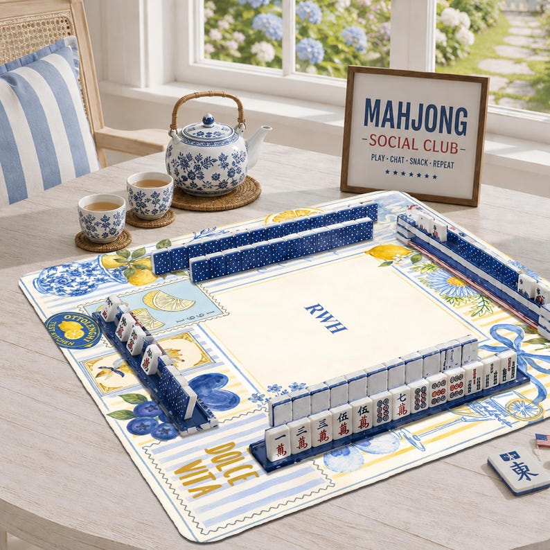 May include: A mahjong set with blue and white tiles arranged on a patterned mat featuring lemons, blueberries, and the text "DOLCE VITA" and "OTTOLENGHI". A blue and white teapot, two matching cups, and a sign reading "MAHJONG SOCIAL CLUB" are present.