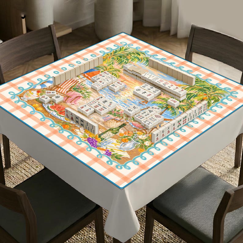 Op de afbeelding: Een vierkant tafelkleed met een strandtafereel met palmbomen, een ligstoel en een picknickmand, omrand door een licht oranje ruitpatroon en een blauwgroene geschulpte lijn. Het tafelkleed is bedekt met witte mahjongtegels in diverse opstellingen.