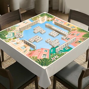 Puede incluir: Un mantel cuadrado para mesa de mahjong con una colorida ilustraci&oacute;n de edificios de estilo mediterr&aacute;neo, follaje verde exuberante y flores rosas. La superficie est&aacute; dispuesta con varios juegos de fichas de mahjong blancas.