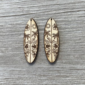 May include: Two wooden surfboard-shaped earrings with etched tropical designs. Each earring features a sun, palm trees, flowers, and waves. The earrings are light brown and have a small hole at the top for hanging.