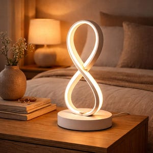 Könnte beinhalten: Eine weiße LED-Tischlampe mit Unendlichkeitszeichen-Design. Die Lampe steht auf einem runden Sockel und strahlt ein warmes Licht aus. Sie steht auf einem Nachttisch aus Holz neben einer Vase und Büchern.