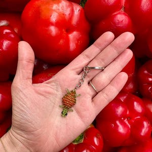 May include: A beaded turtle keychain with orange and green beads, held against a backdrop of bright red bell peppers. The keychain has a silver chain and ring.