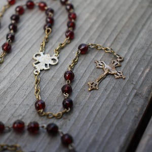 Può includere: Un rosario con perle sferiche rosso scuro traslucide collegate da una catena in metallo color ottone. Il pezzo include un elemento di giunzione centrale in metallo argentato e un crocifisso dettagliato in metallo argentato all'estremità.
