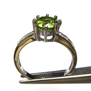 May include: A silver ring with a green gemstone set in a prong setting. The ring has a band of small clear stones.