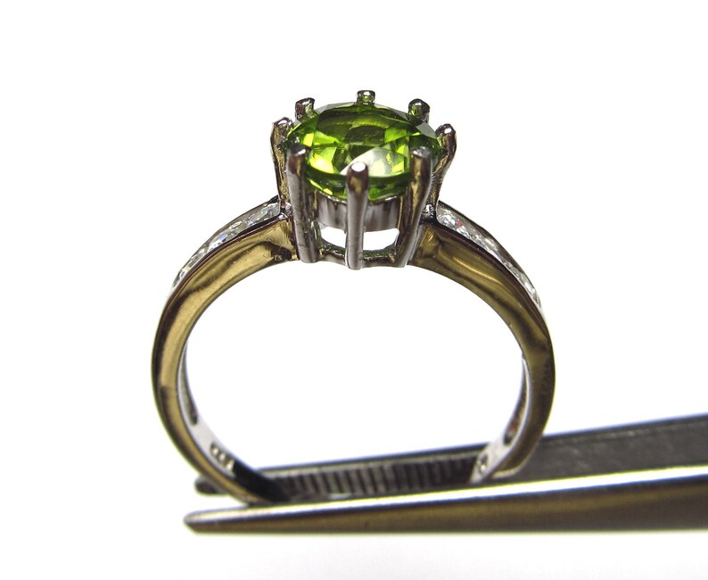May include: A silver ring with a green gemstone set in a prong setting. The ring has a band of small clear stones.