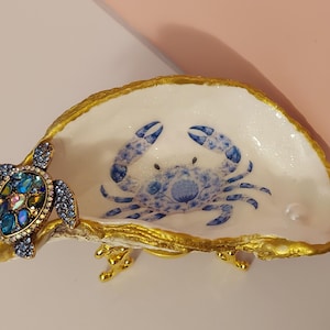May include: A decorative oyster shell dish featuring a hand-painted blue crab design on the interior, with a gold-painted rim and a gold-toned metal stand. A jeweled sea turtle ornament with blue and iridescent crystals is attached to the side.