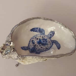 Op de afbeelding: Een decoratieve oesterschelpschaal met een blauw-witte, bloemrijk gedessineerde zeeschildpadillustratie aan de binnenzijde. De rand van de schelp is zilverkleurig geverfd en versierd met een helder gefacetteerd kristal en een kleine witte parel.