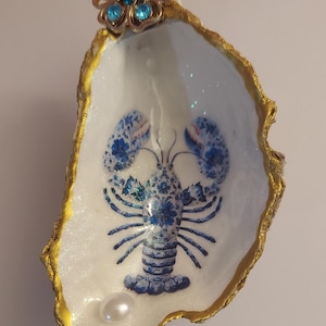 Pode incluir: Uma concha de ostra decorativa com borda dourada. O interior apresenta um desenho de lagosta azul e branco e uma pérola. Um adorno em forma de flor com pedras preciosas azuis adorna a parte superior.