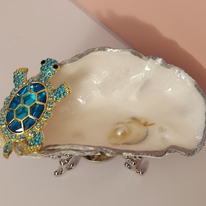 May include: A decorative trinket dish made from a white shell with a silver-toned rim and three small silver legs. A gold-toned turtle figurine embellished with bright blue crystals and a central blue glass piece is attached to the side of the shell.