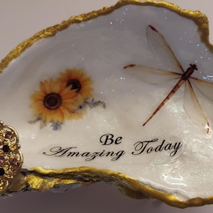 Puede incluir: Un cuenco decorativo de concha de ostra con borde dorado. El cuenco presenta un diseño de libélula y girasol, con las palabras "Be Amazing Today". Lleva un adorno de joyas doradas y moradas.