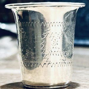 May include: A silver-colored cup with a flared rim and etched geometric patterns. The cup's surface reflects light, highlighting the intricate designs. The cup appears to be made of metal and has a smooth, polished finish.