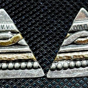 May include: A pair of silver-toned triangle earrings with gold and textured accents. The earrings have a layered design with a beaded bottom edge and a geometric pattern. The background is a black, textured surface.