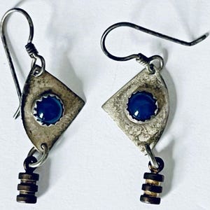 May include: A pair of dangle earrings featuring a triangular silver-toned metal plate with a textured finish and a central circular blue cabochon. Each earring hangs from a curved wire hook and includes a small, dark, three-tiered cylindrical charm.