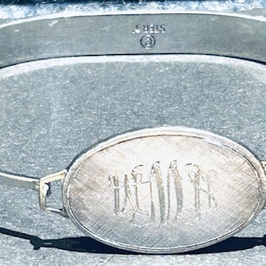 Peut inclure: Un bracelet jonc argenté avec une pièce maîtresse ovale. La pièce maîtresse présente un motif gravé. Le bracelet est marqué "STERLING". Le bracelet est un bijou simple et élégant.