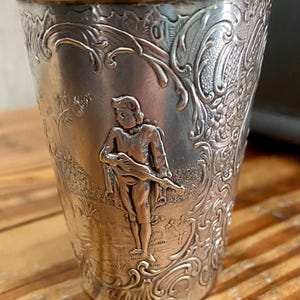 May include: A silver-toned metal cup featuring an embossed scene of a person playing a stringed instrument in a landscape. The exterior surface is decorated with intricate scrollwork, floral patterns, and a textured background finish.