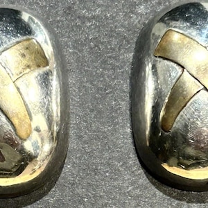 Könnte beinhalten: Ein Paar silberfarbene, ovale Ohrringe mit einem goldfarbenen X-Design. Die Ohrringe haben eine polierte Oberfläche und sind vor einem dunklen Hintergrund abgebildet. Das goldfarbene X ist auf jedem Ohrring zentriert.
